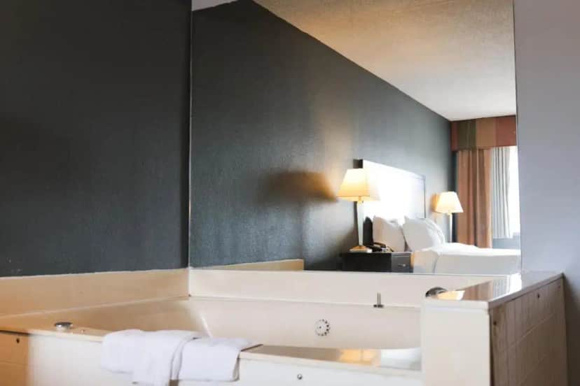a bathroom with a tub and a bed and a mirror at Baymont by Wyndham Flint Airport North in Flint